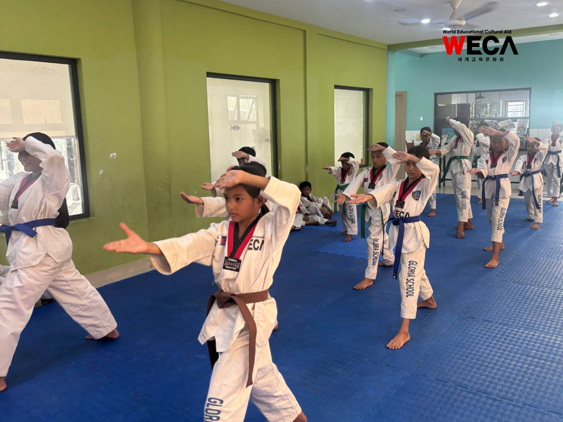 캄보디아 프놈펜 안짠 글로리아 WECA 태권도 교실에서 사범의 지도 아래 24명의 아이들이 도복을 입고 기본 동작을 연습하는 모습